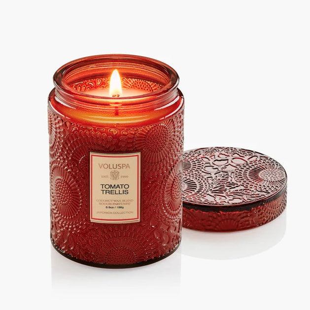 A red embossed glass jar candle with a lit flame, shown next to its matching patterned lid on a clean white background. The label reads Voluspa Tomato Trellis.