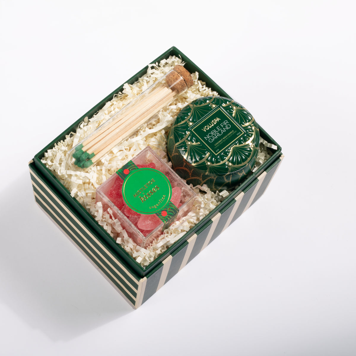 A curated gift box set on a white background, containing a green and white striped box filled with crinkle paper. Inside are a tin of scented soap, a small clear package with a green lid, wooden matchsticks with green tips, and a green decorative tag.