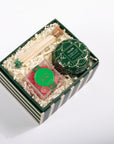 A curated gift box set on a white background, containing a green and white striped box filled with crinkle paper. Inside are a tin of scented soap, a small clear package with a green lid, wooden matchsticks with green tips, and a green decorative tag.
