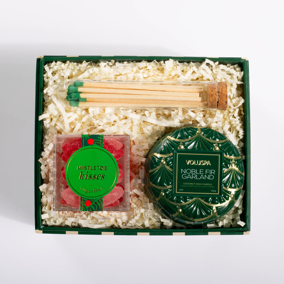 A curated gift box set containing a green and gold Voluspa Noble Fir Garland tin, a square box of red mistletoe kisses, a glass of green-tipped matches, and white shredded paper inside a dark green gift box, photographed from a top-down angle on a white background.