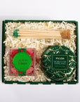 A curated gift box set containing a green and gold Voluspa Noble Fir Garland tin, a square box of red mistletoe kisses, a glass of green-tipped matches, and white shredded paper inside a dark green gift box, photographed from a top-down angle on a white background.