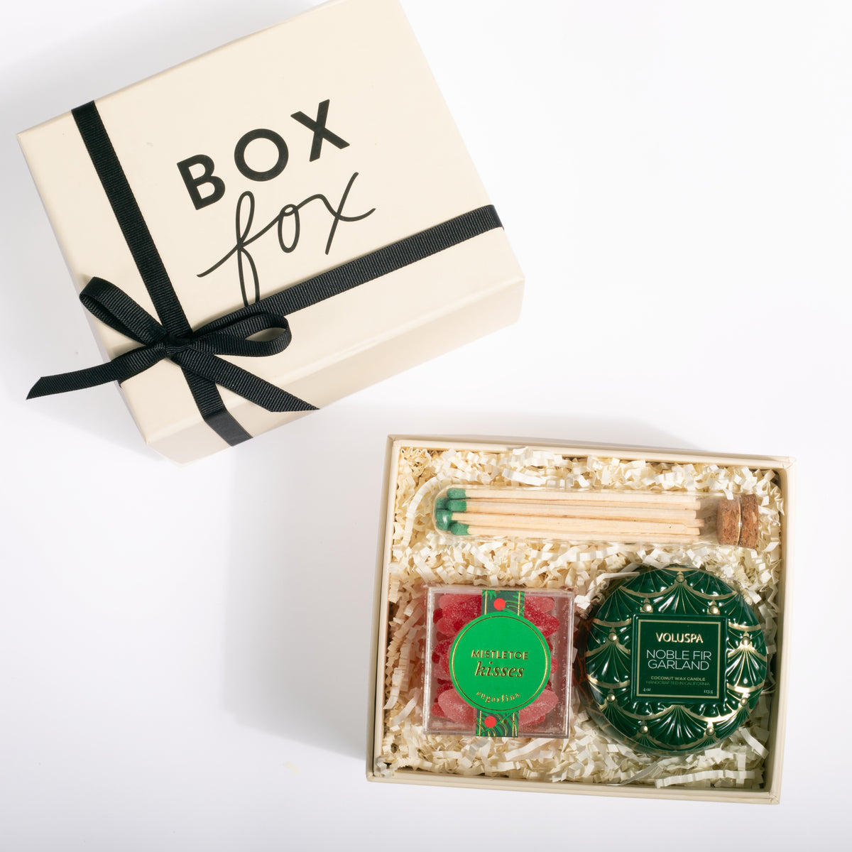 A gift box set from Box Fox, featuring a cream-colored box with a black ribbon and bow next to an open box filled with shredded paper and containing a green tea and vanilla extract blend, wooden matches, and a green decorative tin.
