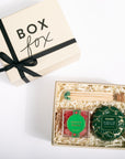 A gift box set from Box Fox, featuring a cream-colored box with a black ribbon and bow next to an open box filled with shredded paper and containing a green tea and vanilla extract blend, wooden matches, and a green decorative tin.