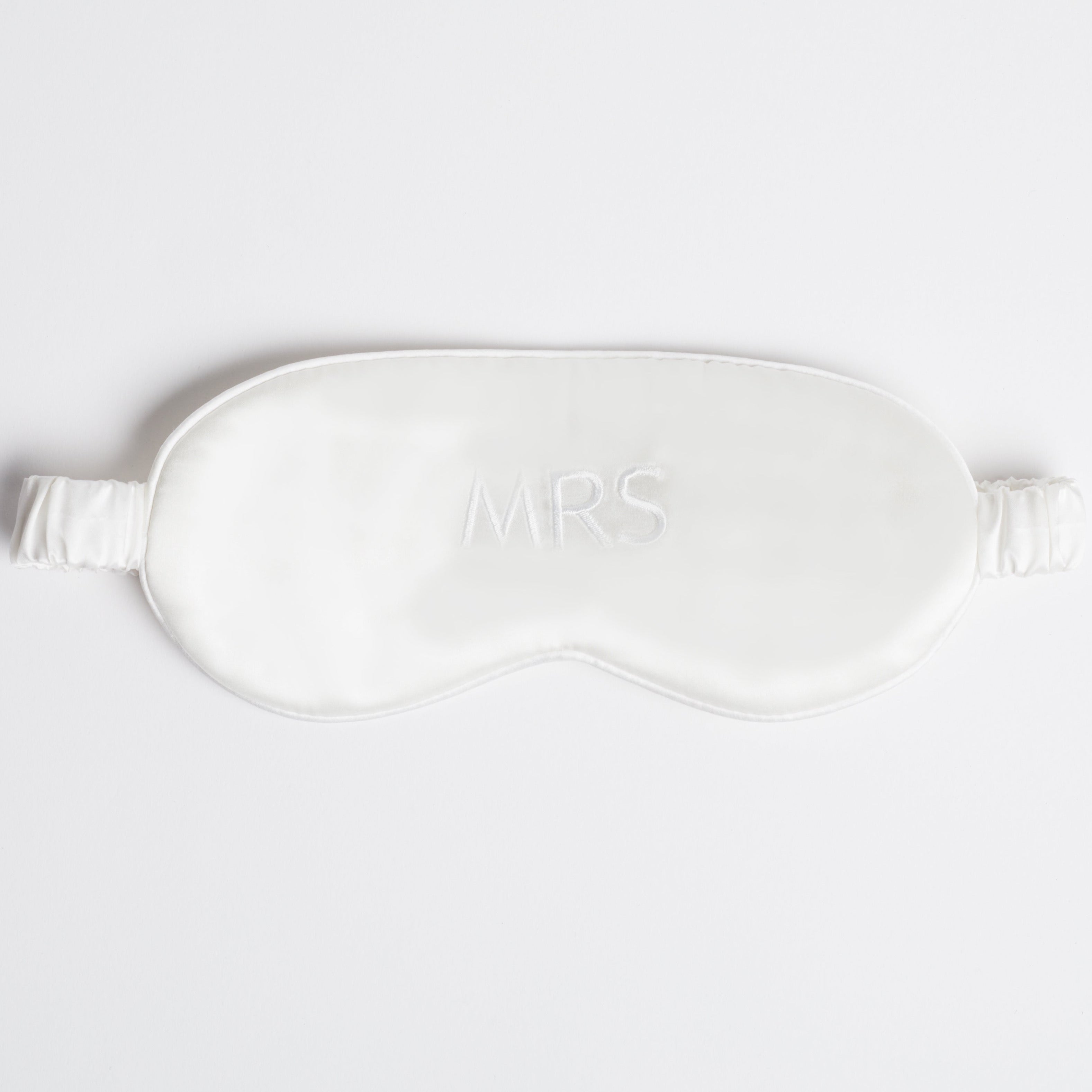 White silk eye mask with "MRS" written across it, lying on a white background.