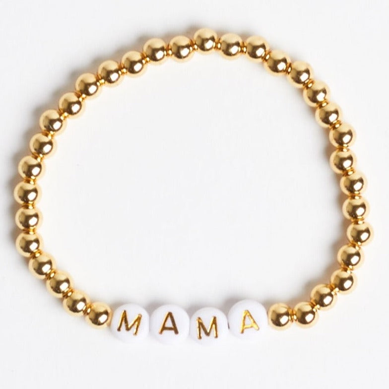 A gold beaded stretch bracelet with white beads that have gold text reading "M" "A" "M" "A" to spell out "MAMA." Photographed against a white background.
