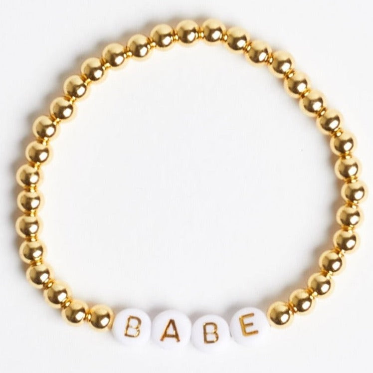 A gold beaded stretch bracelet with white beads that have gold text reading &quot;B&quot; &quot;A&quot; &quot;B&quot; &quot;E&quot; to spell out &quot;BABE.&quot; Photographed against a white background.