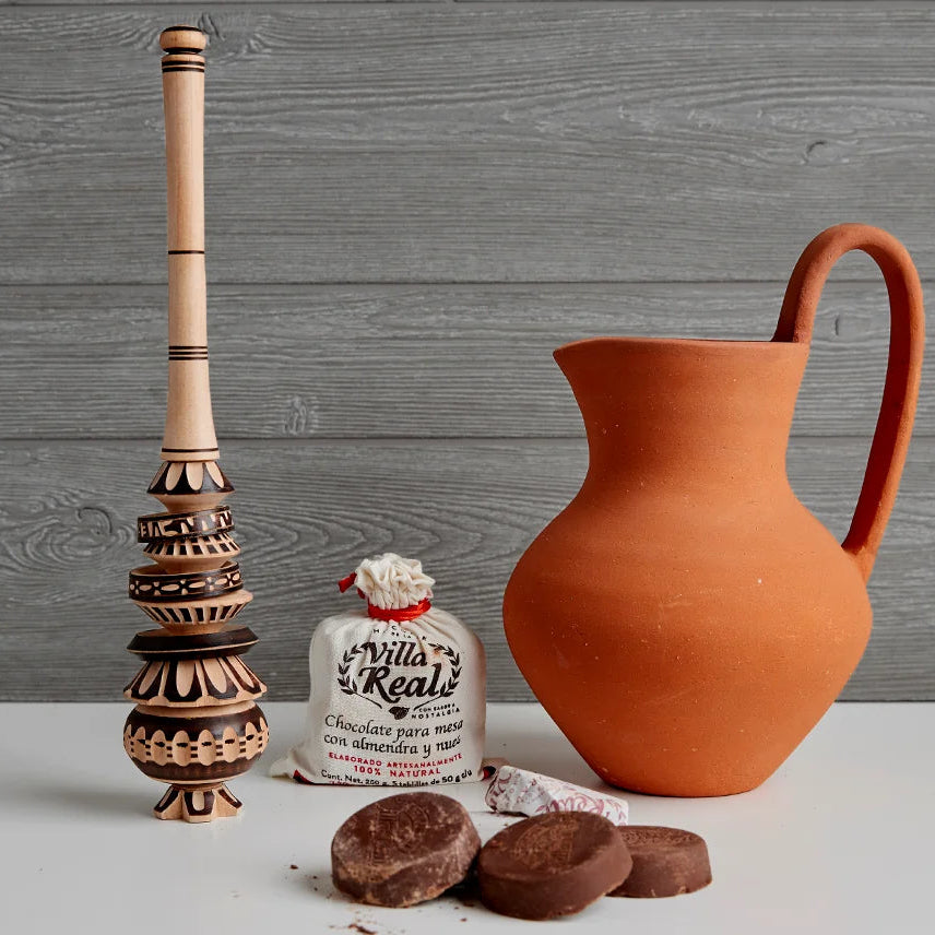 Terracotta pitcher, wooden pestle, and a bottle of Villa Real's chocolate cream on a wooden surface.