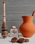 Terracotta pitcher, wooden pestle, and a bottle of Villa Real's chocolate cream on a wooden surface.