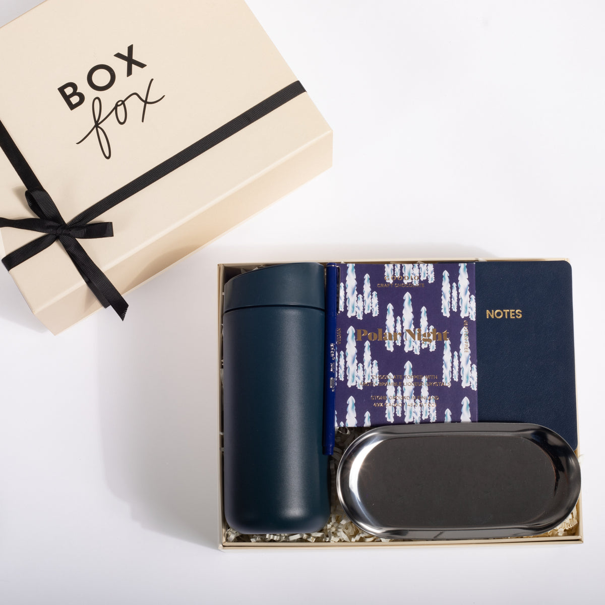 A gift set from Box Fox on a white background, featuring a navy blue insulated tumbler, a navy notebook with a purple patterned cover, a black metal tray, and the beige gift box with a black ribbon.