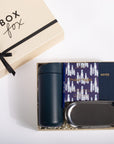 A gift set from Box Fox on a white background, featuring a navy blue insulated tumbler, a navy notebook with a purple patterned cover, a black metal tray, and the beige gift box with a black ribbon.