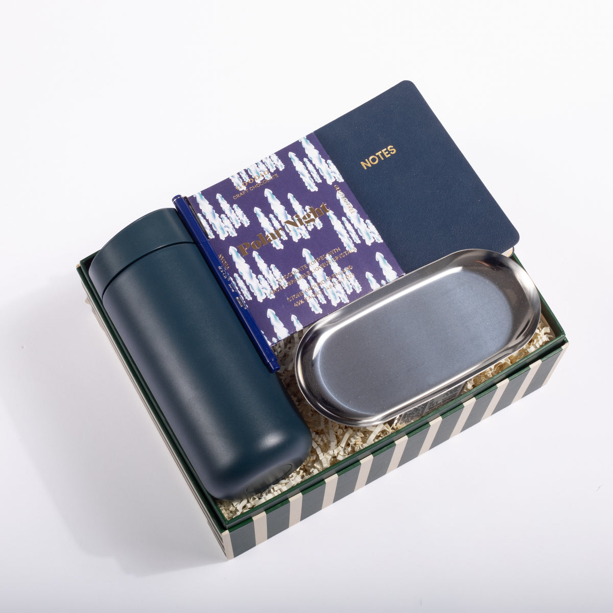 A gift set in a striped box containing a dark teal insulated bottle, a navy notebook with gold foil 'NOTES' text, a patterned purple notebook, and a stainless steel tray on a white background.