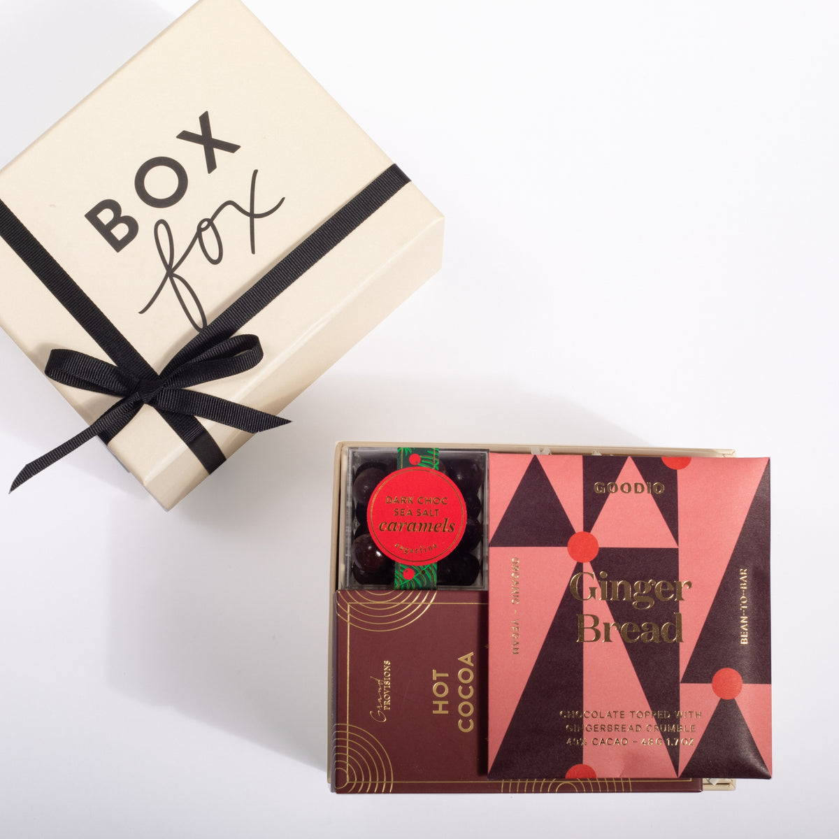 A gift box set from Goodies featuring a cream-colored 'Box fox' box with a black ribbon, alongside an open gift box containing chocolate gingerbread cookies, hot cocoa, and a small jar of caramel.