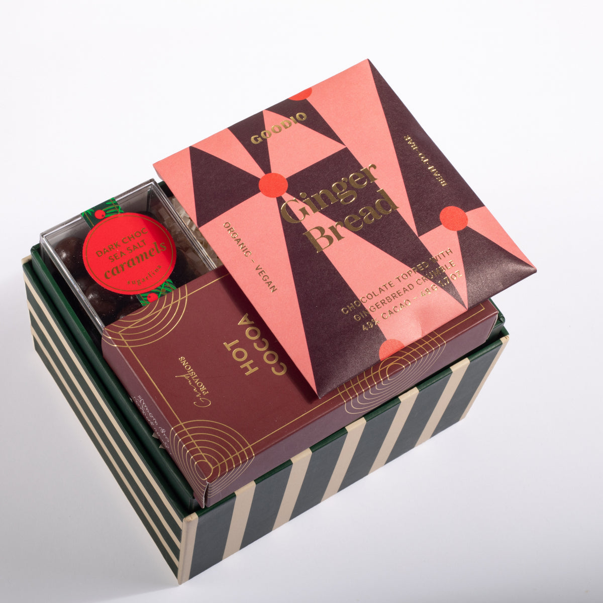 A gourmet gift box set containing a gingerbread cookie, a hot cocoa box, and a small caramel candy, presented in a striped box on a white background.