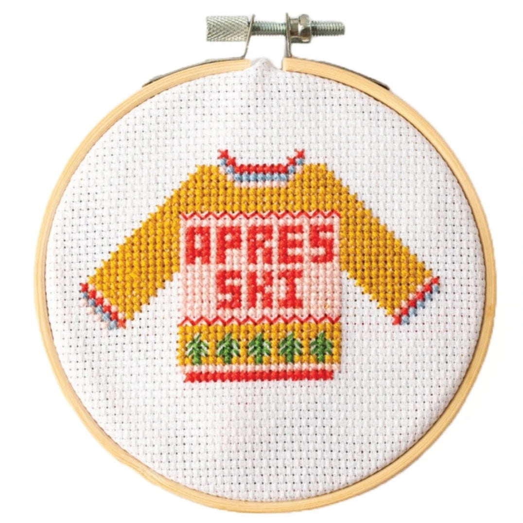 This image shows a cross-stitch embroidery hoop featuring a cozy, colorful sweater design with the text "APRÈS SKI" stitched across the front. The sweater is depicted in warm hues, with mustard yellow sleeves and a pink torso. It features decorative patterns at the cuffs, hem, and neckline, as well as a row of small green tree-like shapes near the bottom, reminiscent of festive motifs. The embroidery is framed within a wooden hoop, giving it a charming handcrafted look, perfect for winter or ski-theme.