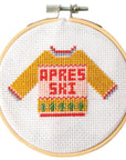 This image shows a cross-stitch embroidery hoop featuring a cozy, colorful sweater design with the text "APRÈS SKI" stitched across the front. The sweater is depicted in warm hues, with mustard yellow sleeves and a pink torso. It features decorative patterns at the cuffs, hem, and neckline, as well as a row of small green tree-like shapes near the bottom, reminiscent of festive motifs. The embroidery is framed within a wooden hoop, giving it a charming handcrafted look, perfect for winter or ski-theme.