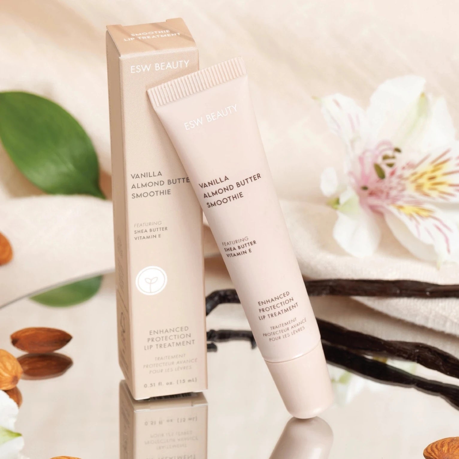 The image displays a tube and box of "ESW Beauty Vanilla Almond Butter Smoothie," described as an enhanced protection lip treatment. The packaging highlights key ingredients such as shea butter and vitamin E, known for their nourishing properties. The tube and box are presented on a reflective surface surrounded by natural elements, including almonds, vanilla beans, green leaves, and delicate white flowers, evoking a sense of freshness and natural care. There is soft beige and cream tones of the packaging.