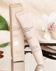 The image displays a tube and box of "ESW Beauty Vanilla Almond Butter Smoothie," described as an enhanced protection lip treatment. The packaging highlights key ingredients such as shea butter and vitamin E, known for their nourishing properties. The tube and box are presented on a reflective surface surrounded by natural elements, including almonds, vanilla beans, green leaves, and delicate white flowers, evoking a sense of freshness and natural care. There is soft beige and cream tones of the packaging.