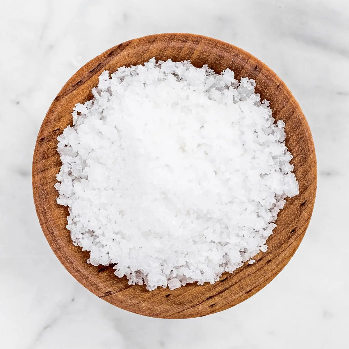 Coarse white sea salt crystals piled in a round wooden bowl on a light marble surface.