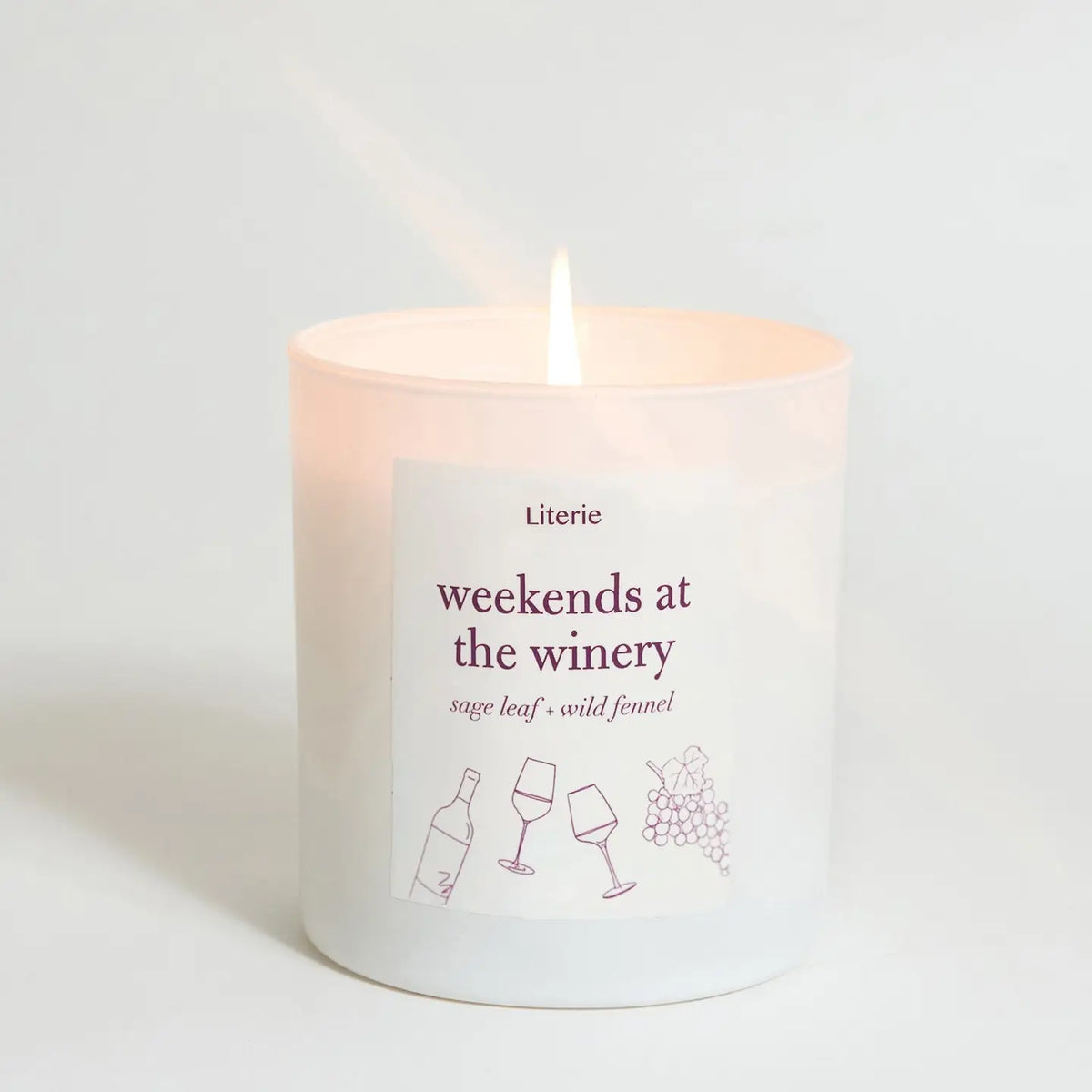 A lit white glass candle with a single flame, featuring a white label that reads 'Literie weekends at the winery' with wine bottle and glass illustrations, set against a clean white background.