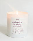 A lit white glass candle with a single flame, featuring a white label that reads 'Literie weekends at the winery' with wine bottle and glass illustrations, set against a clean white background.
