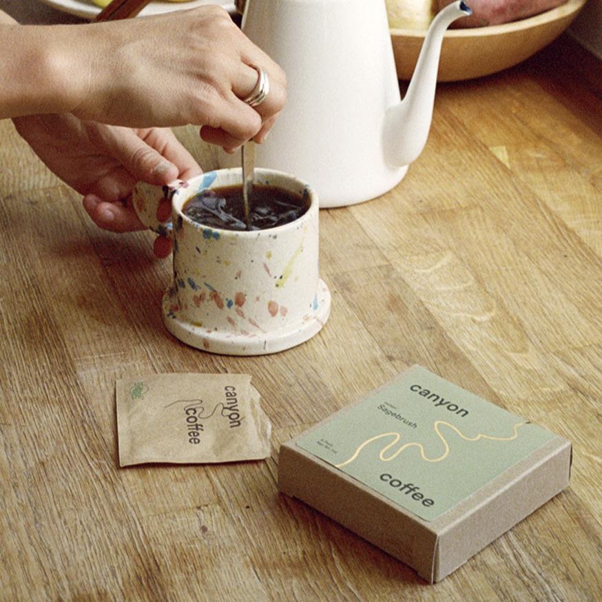 sage green instant coffee box next to hand stirring spoon in terracotta coffee mug