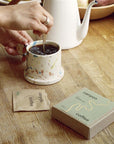 sage green instant coffee box next to hand stirring spoon in terracotta coffee mug