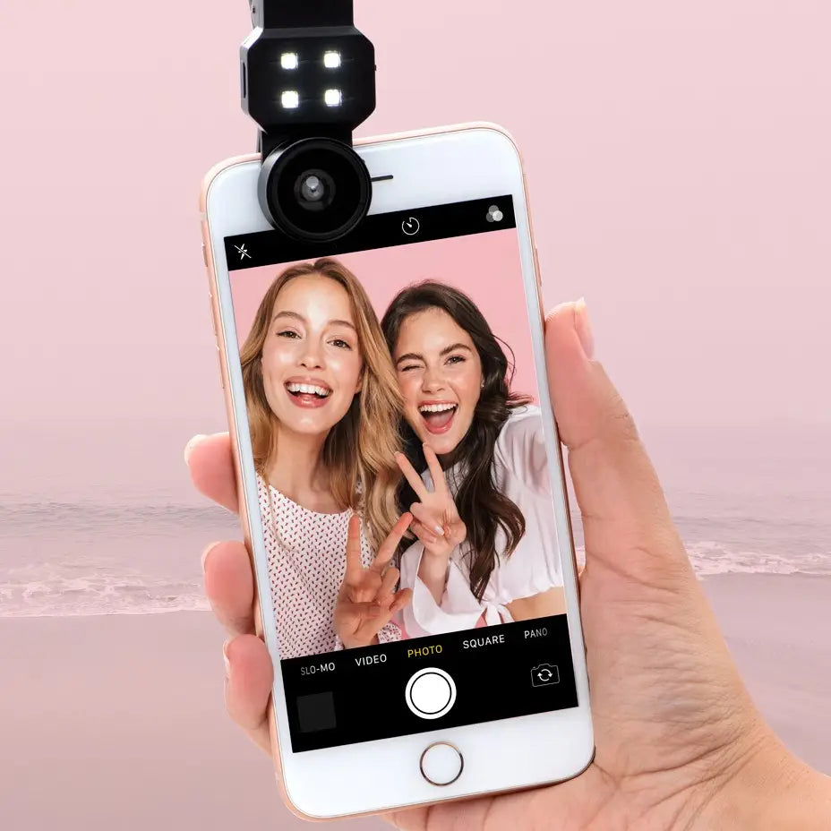 Hand holding a smartphone with a selfie light attached, showing a photo of two people on the screen.