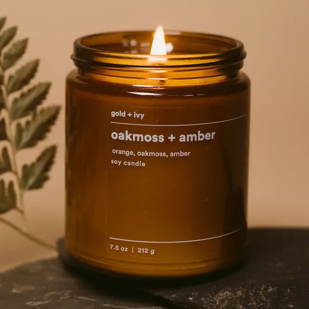 Brown candle labeled 'oakmoss + amber' with a flame on a dark surface.