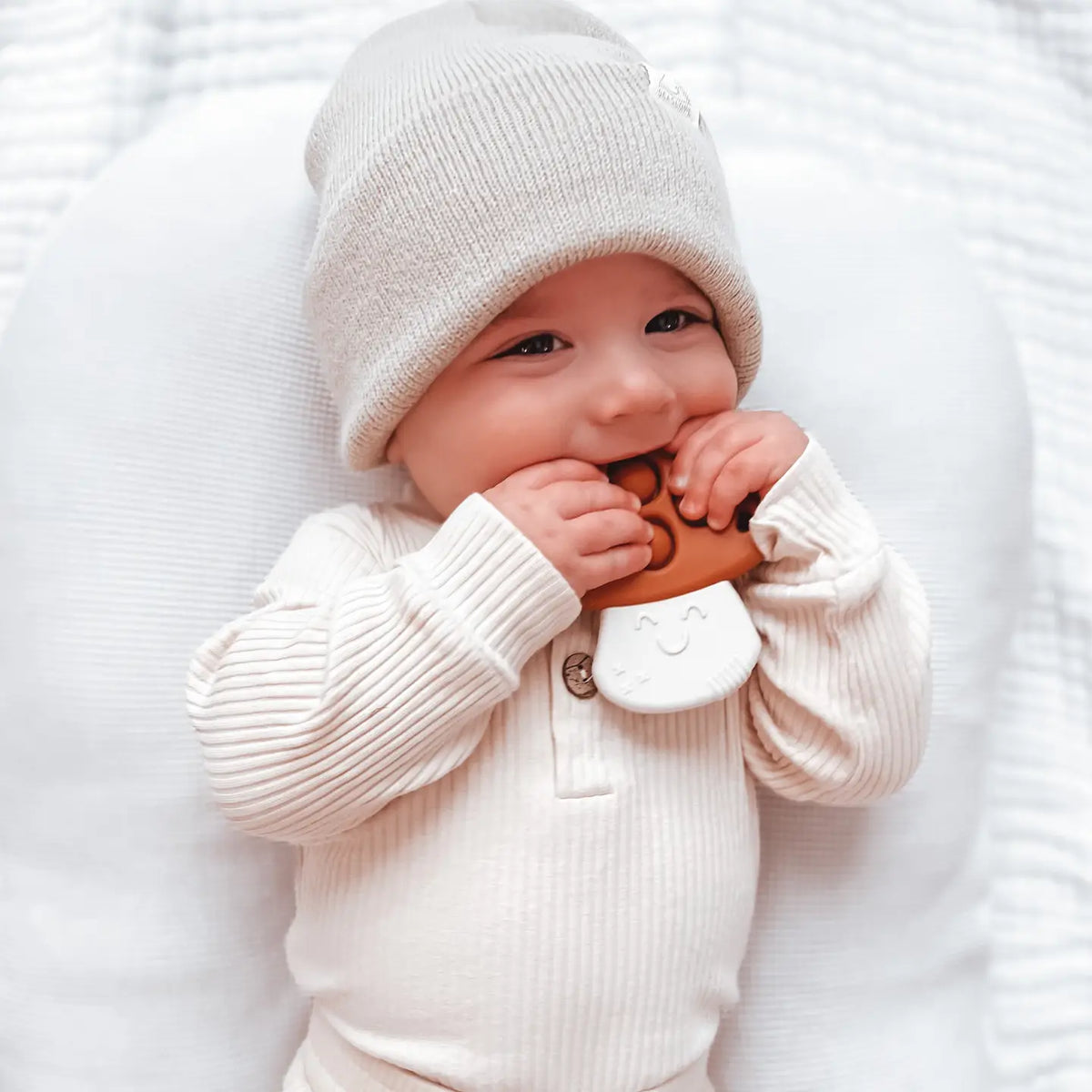 A baby wearing a cream ribbed knit hat and matching long-sleeve top lies on a white textured blanket, chewing on a brown bear-shaped teething toy with a white belly detail.