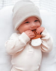 A baby wearing a cream ribbed knit hat and matching long-sleeve top lies on a white textured blanket, chewing on a brown bear-shaped teething toy with a white belly detail.