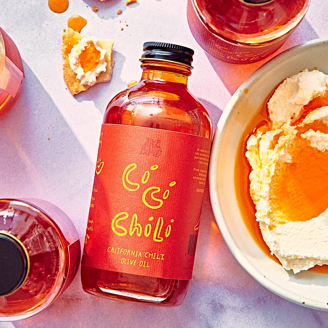 Bottle of Có Có Chólo olive oil with a bowl of hummus and olive oil glasses on a light background