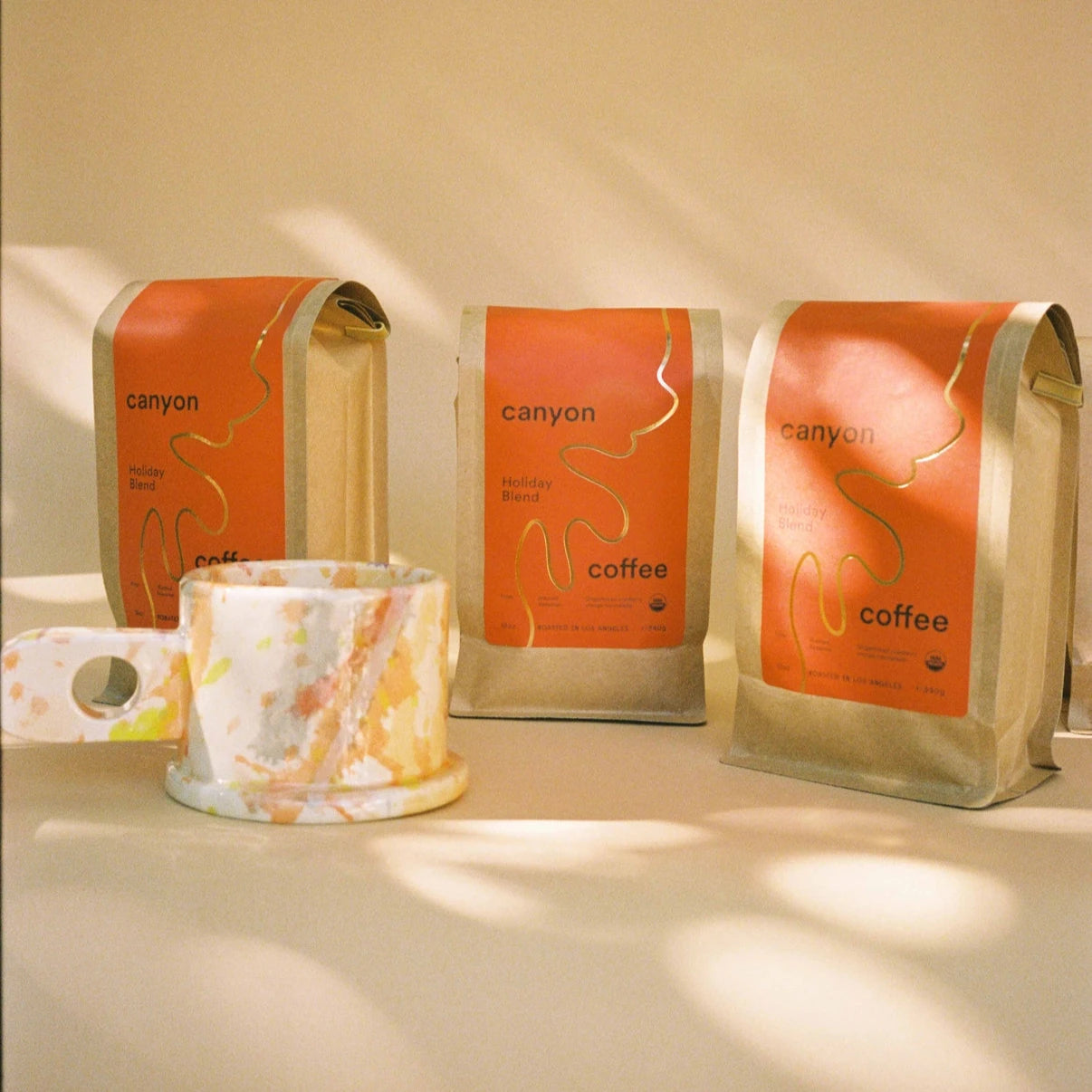 A collection of three Canyon Coffee Holiday Blend bags displayed upright with a colorful, speckled ceramic mug beside them. The background is softly lit, adding a cozy and artistic vibe.

