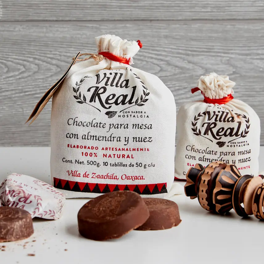 Two white drawstring gift bags of Villa Real chocolate conchito are displayed on a white surface with chocolate pieces and wooden spoons in front, against a gray wood-textured background.