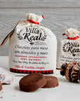 Two white drawstring gift bags of Villa Real chocolate conchito are displayed on a white surface with chocolate pieces and wooden spoons in front, against a gray wood-textured background.