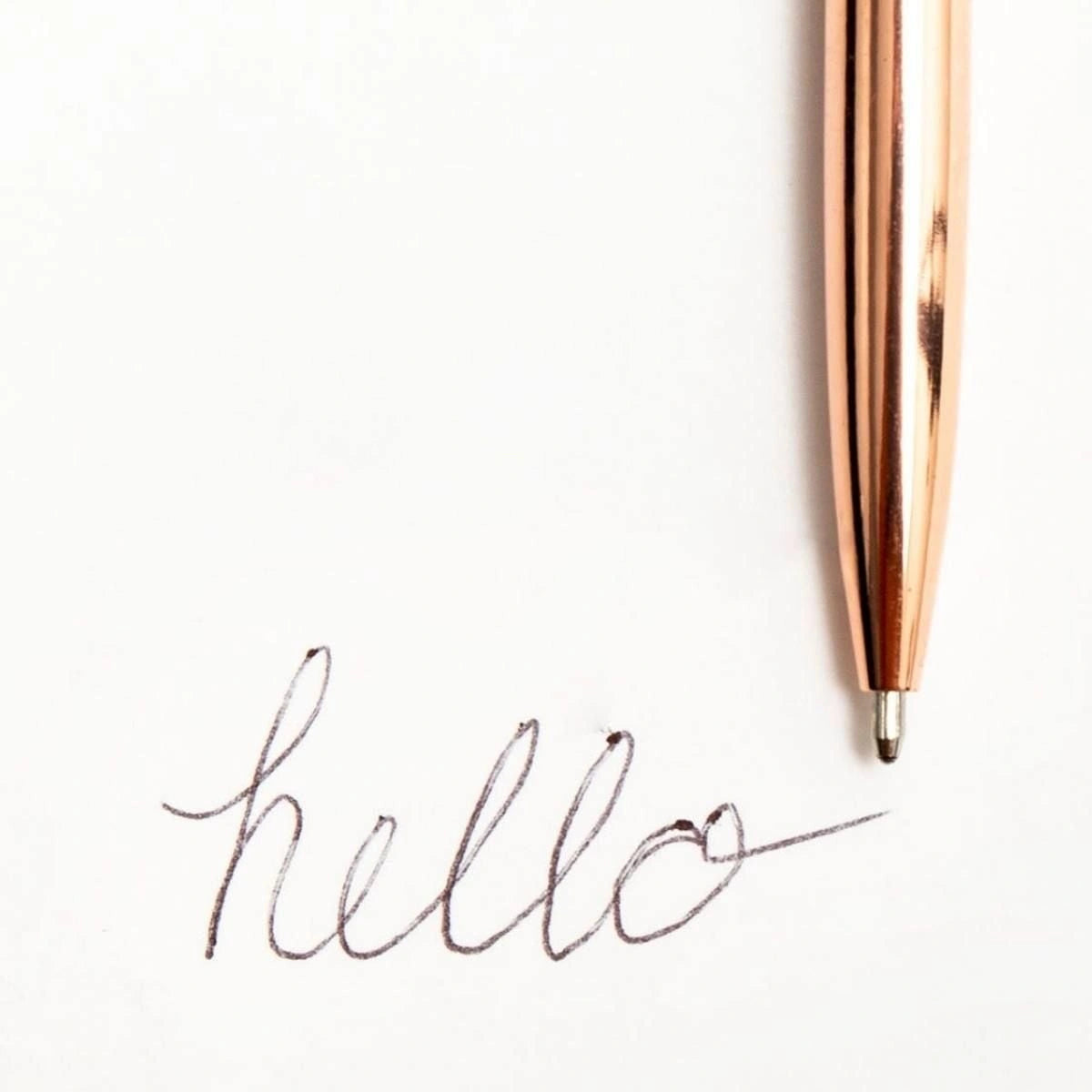 A close-up of a metallic rose gold pen resting on a white sheet of paper next to a cursive 'hello' signature.