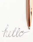 A close-up of a metallic rose gold pen resting on a white sheet of paper next to a cursive 'hello' signature.