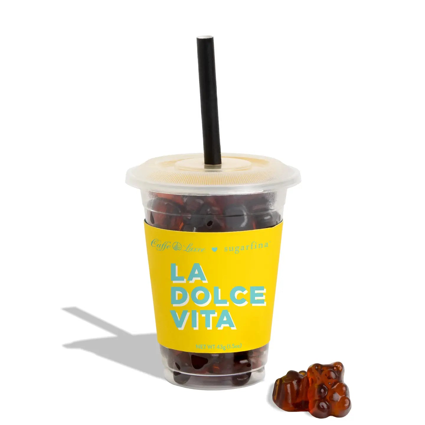 A clear plastic cup with a translucent lid and black straw, filled with dark gummy candies and wrapped in a bright yellow sleeve printed with the words 'LA DOLCE VITA'. A small cluster of brown gummy bears sits beside the cup on a white background.