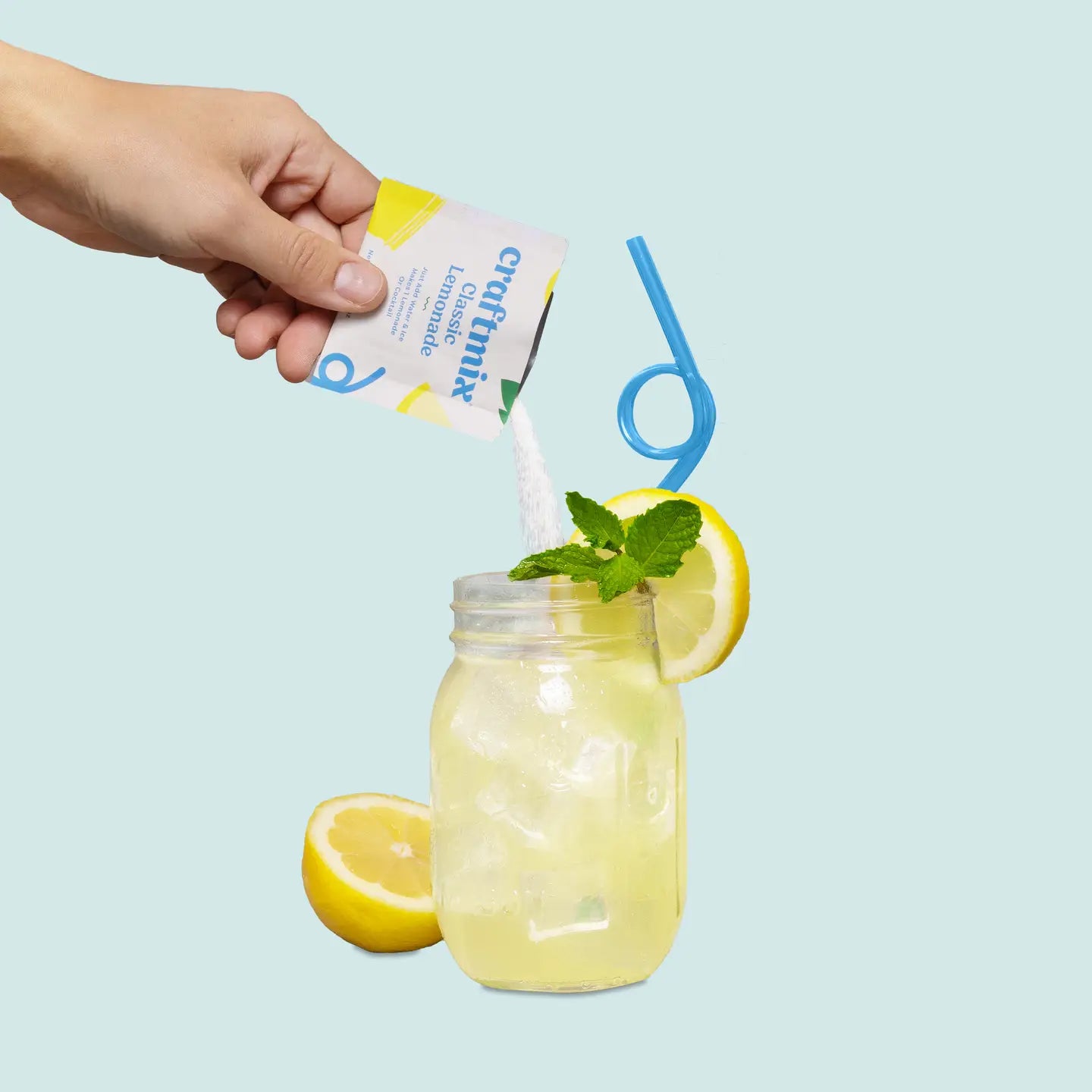Hand pouring a drink mix into a glass with lemon slices and a straw on a light blue background