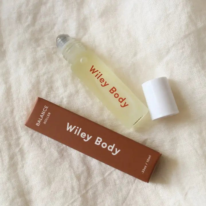 A small clear glass roller bottle labeled 'Wiley Body' with a white cap, placed beside a brown rectangular box also branded 'Wiley Body' on a textured off-white fabric background.