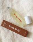 A small clear glass roller bottle labeled 'Wiley Body' with a white cap, placed beside a brown rectangular box also branded 'Wiley Body' on a textured off-white fabric background.