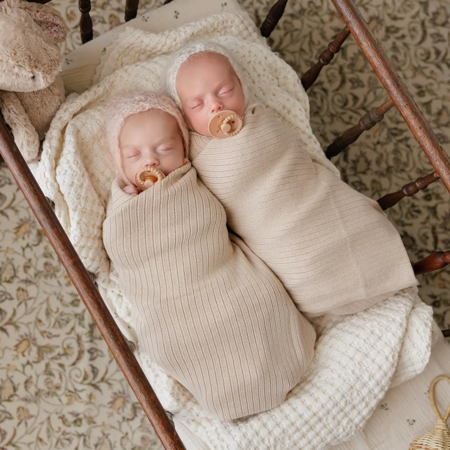 Two newborn babies swaddled in beige wraps with pacifiers, lying on a wooden crib.