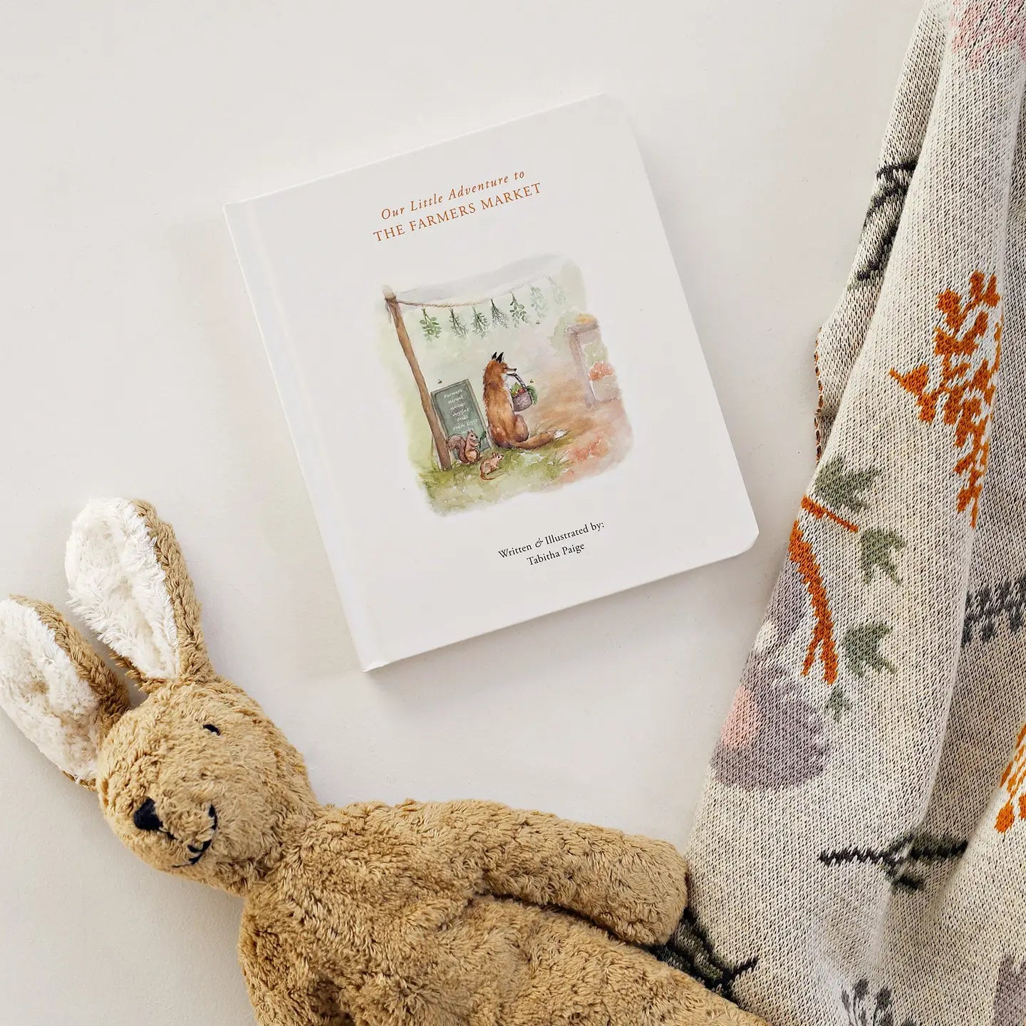 Children's book titled 'Our Little Adventure to the Farmer's Market' with a plush rabbit toy and patterned blanket on a light background.