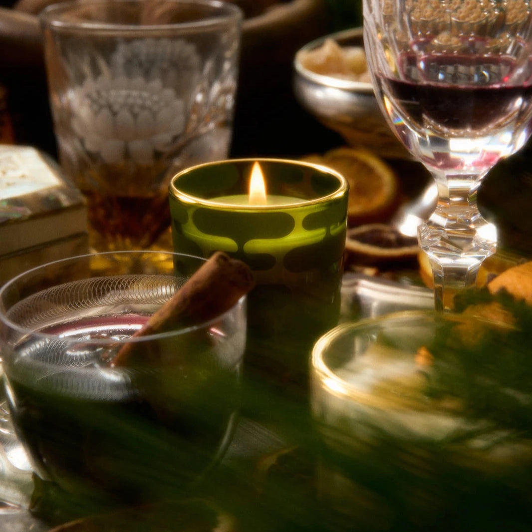 A warm holiday scene with candles, drinks, and seasonal decorations. The focus is on a lit candle in a green patterned glass, surrounded by a glass of mulled wine, crystal glasses of amber and red beverages, and a bowl of walnuts in the background. Fresh greens add to the cozy, festive mood.