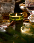 A warm holiday scene with candles, drinks, and seasonal decorations. The focus is on a lit candle in a green patterned glass, surrounded by a glass of mulled wine, crystal glasses of amber and red beverages, and a bowl of walnuts in the background. Fresh greens add to the cozy, festive mood.