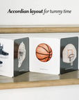 Accordian layout book for tummy time with sports-themed illustrations on a wooden surface.