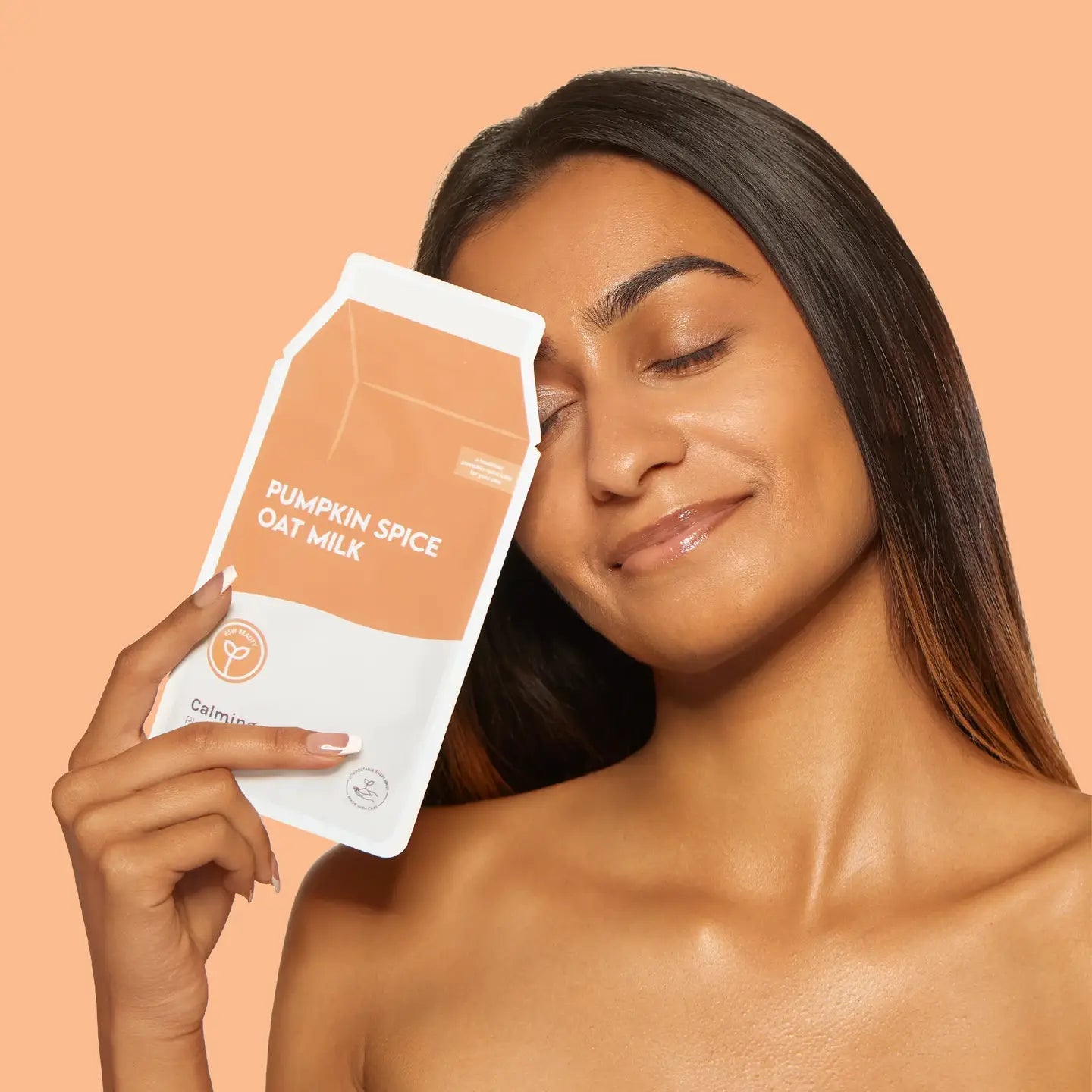 A woman with long dark hair holds a pouch of pumpkin spice oat milk next to her face against a solid peach background.