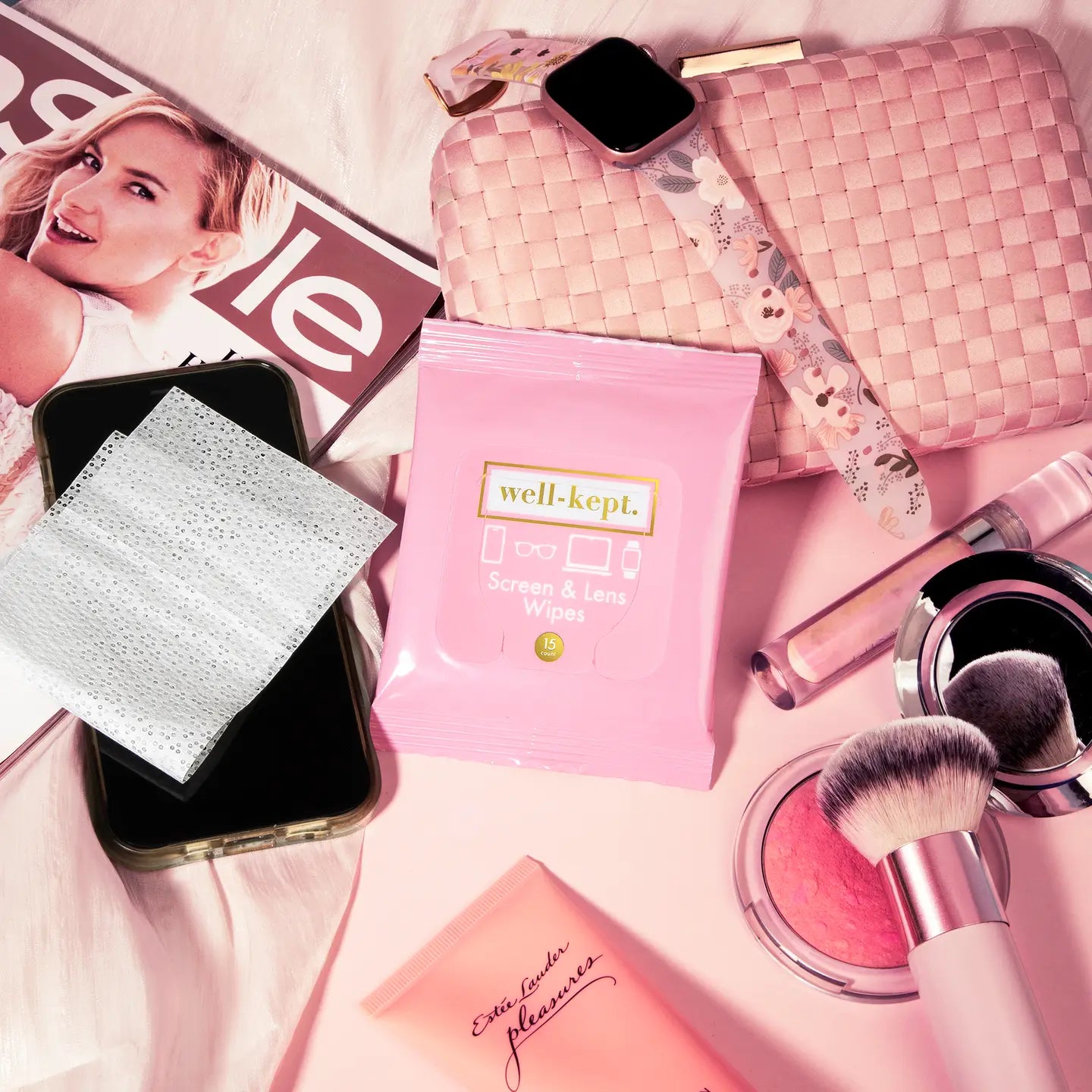Makeup products including brushes, a phone, and a pink bag on a pink surface.