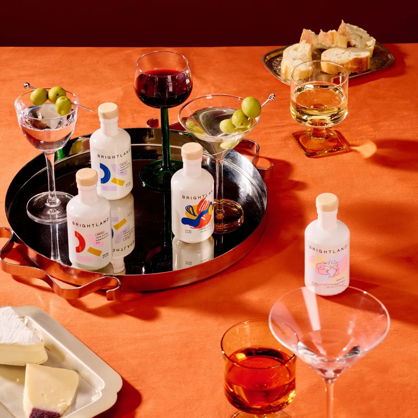 Brightland artisanal spirits bottles arranged on a black serving tray with cocktails, olives, bread, and cheese on an orange tablecloth.