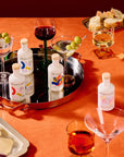 Brightland artisanal spirits bottles arranged on a black serving tray with cocktails, olives, bread, and cheese on an orange tablecloth.