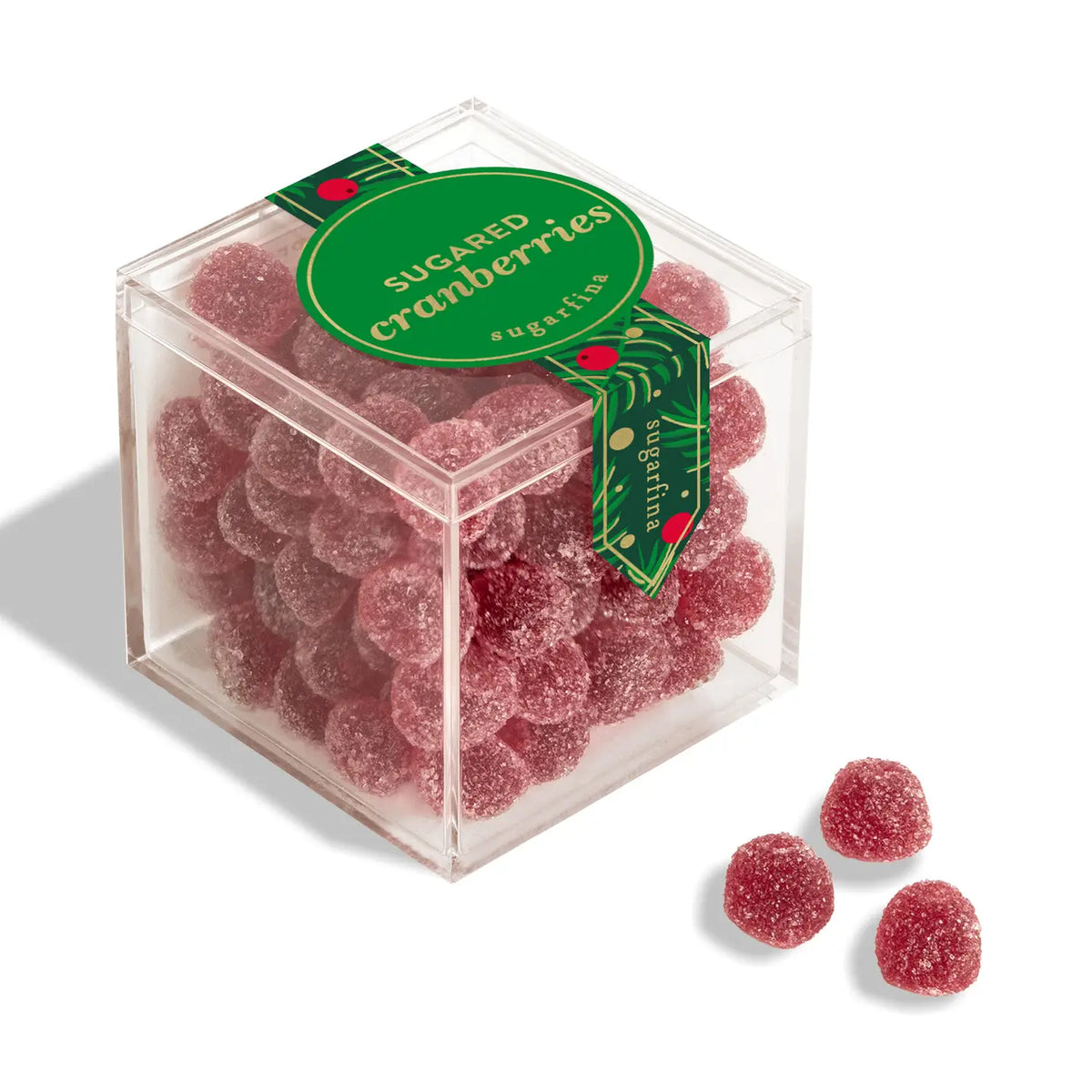 A clear square plastic box filled with round, sugar-coated cranberry candies, with a green circular label reading 'SUGARED cranberries' and a decorative ribbon. Three additional cranberry candies are placed outside the box on a white background.